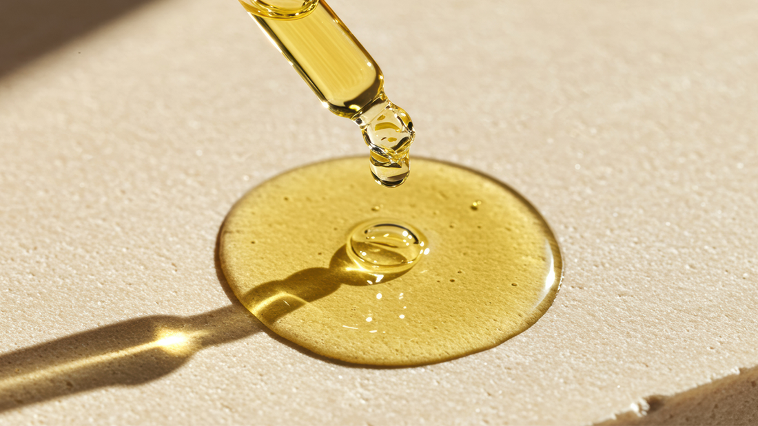 Golden cosmetic oil dripping from a glass pipette onto a soft beige surface, illustrating the texture of nourishing hair and scalp oils