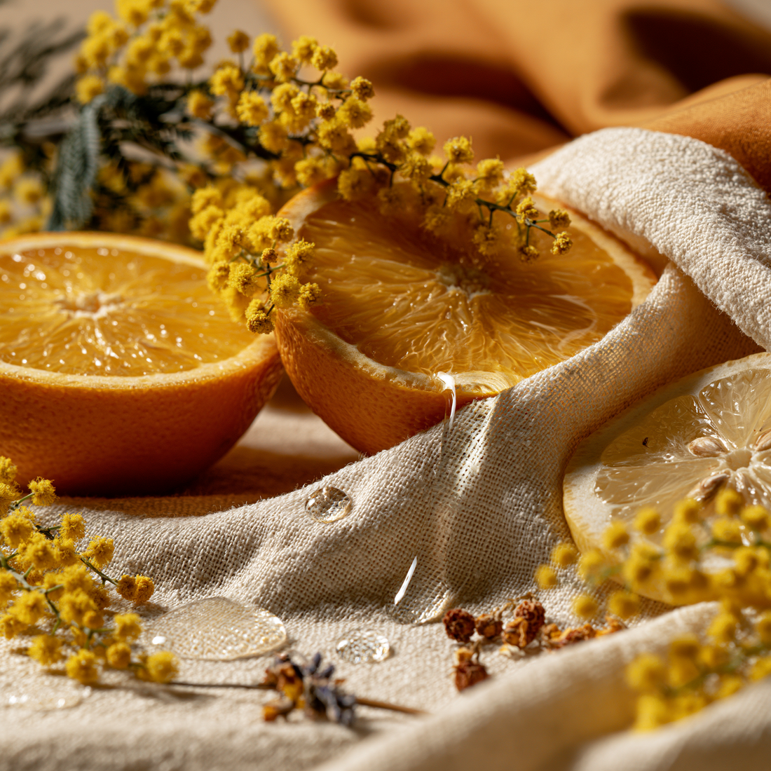 "A halved orange, sliced lemon and small amber glass dropper bottle arranged on cream linen with dried mimosa flowers in soft morning light"