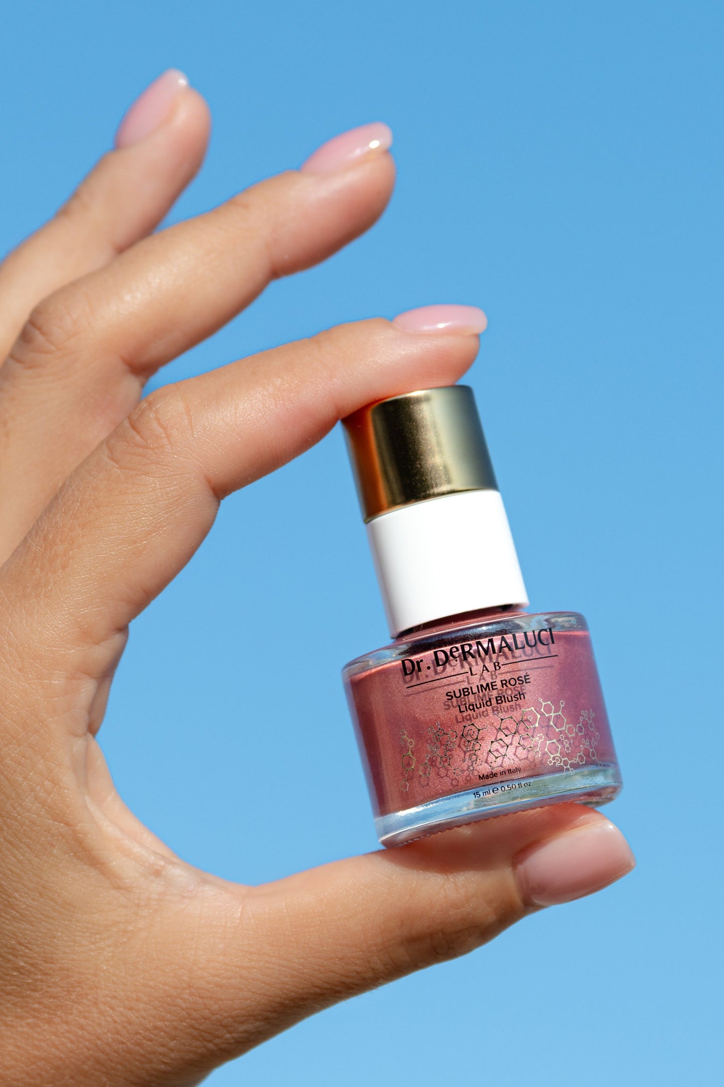 Hand holding a bottle of Dr. Dermaluci Lab Rose Blush against a blue background